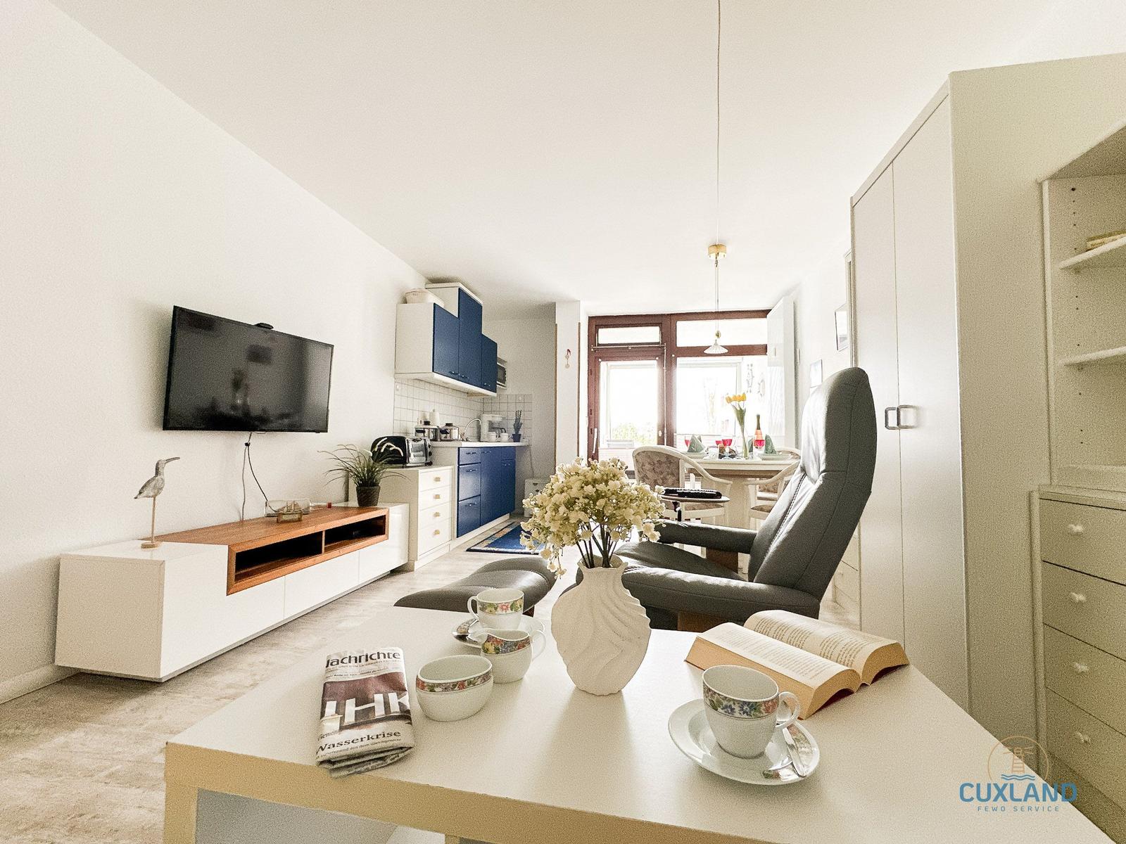 Wohnzimmer mit TV, Küche und Essbereich. Tisch mit Tassen und Blumen.