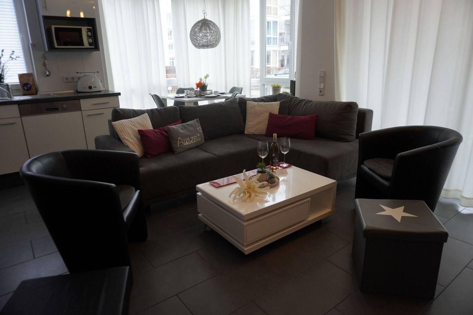 Wohnzimmer mit Sofa, zwei Sesseln und Couchtisch. Küchenbereich im Hintergrund.