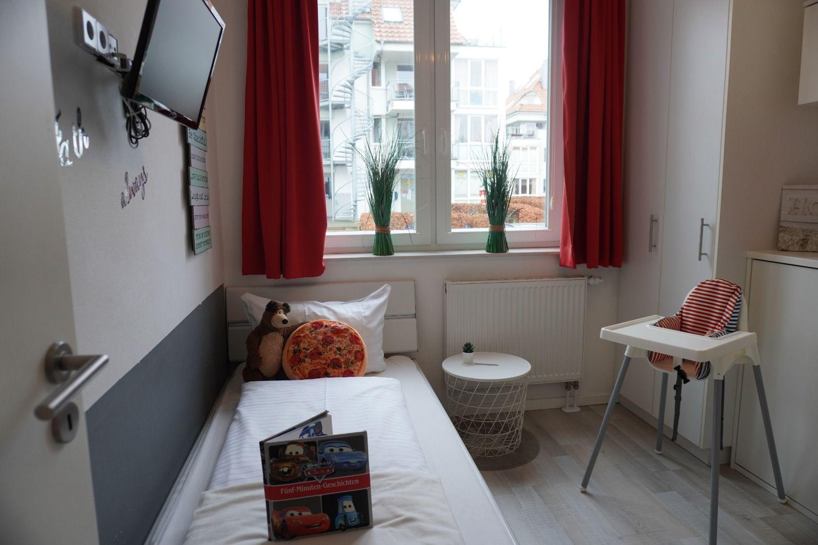 Kinderzimmer mit Bett, Hochstuhl und Fenster mit roten Vorhängen.