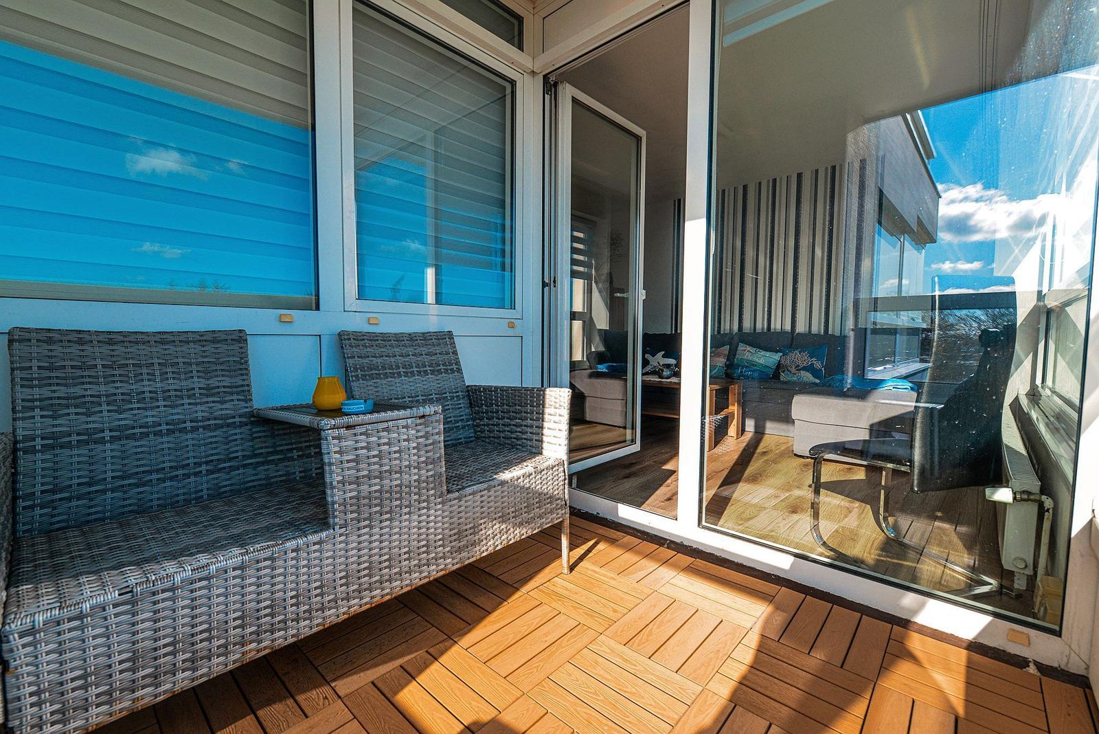 Terrasse mit Rattanmöbeln und Holzboden. Durchgehendes Glas führt ins Wohnzimmer.