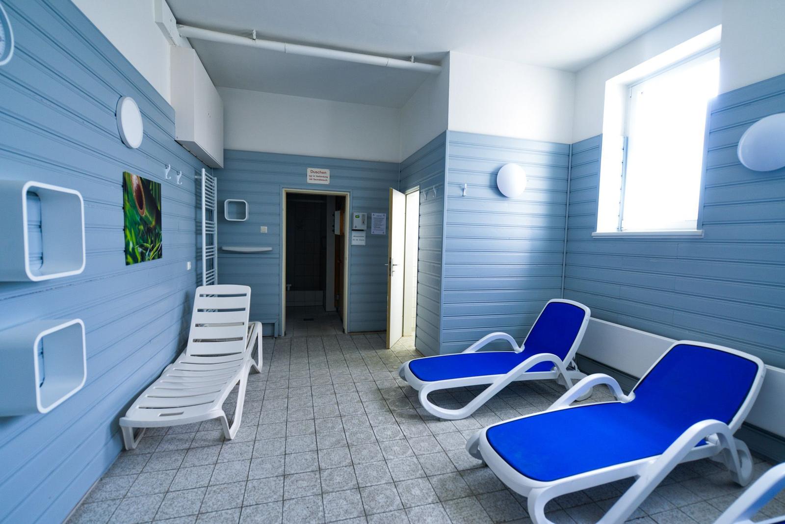 Raum mit blauen Wänden, Liegen und Fenster. Tür führt in Duschenbereich.