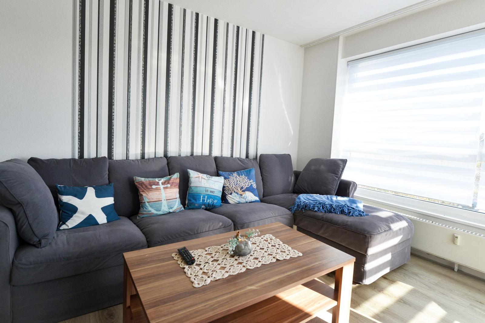 Ein Wohnzimmer mit grauem Sofa, dekorativen Kissen und Holztisch vor großem Fenster.