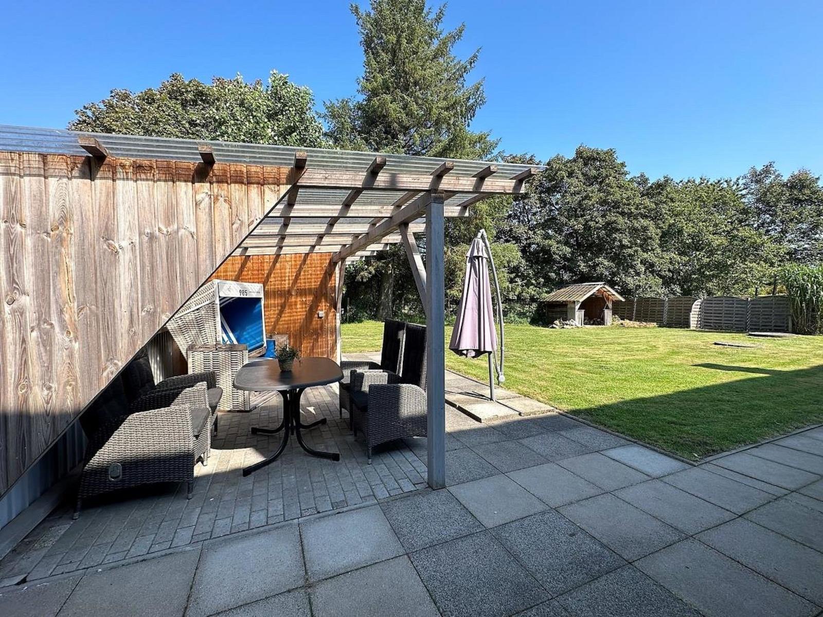 Terrasse mit Sitzgruppe unter Dach, Rasenfläche und Holzhaus im Hintergrund.
