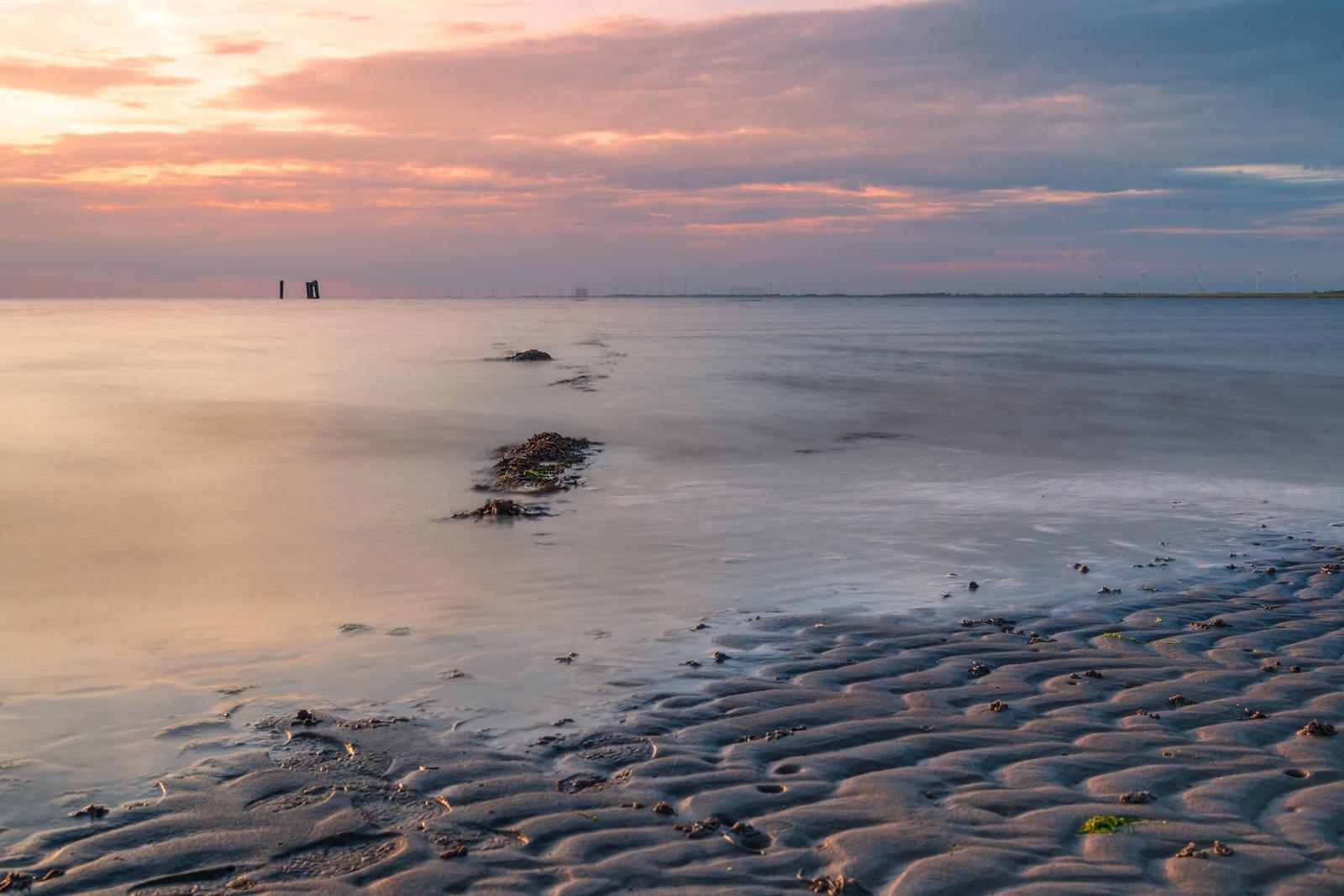Strand mit Wellen und Sandwellen bei Sonnenuntergang mit Windkraftanlagen im Hintergrund