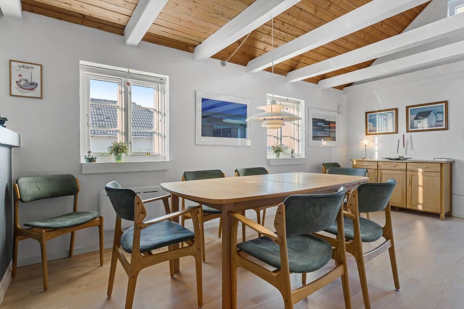 Esszimmer mit Holztisch, Stühlen und Fenstern. Decke mit Holzbalken und Bildern an den Wänden.