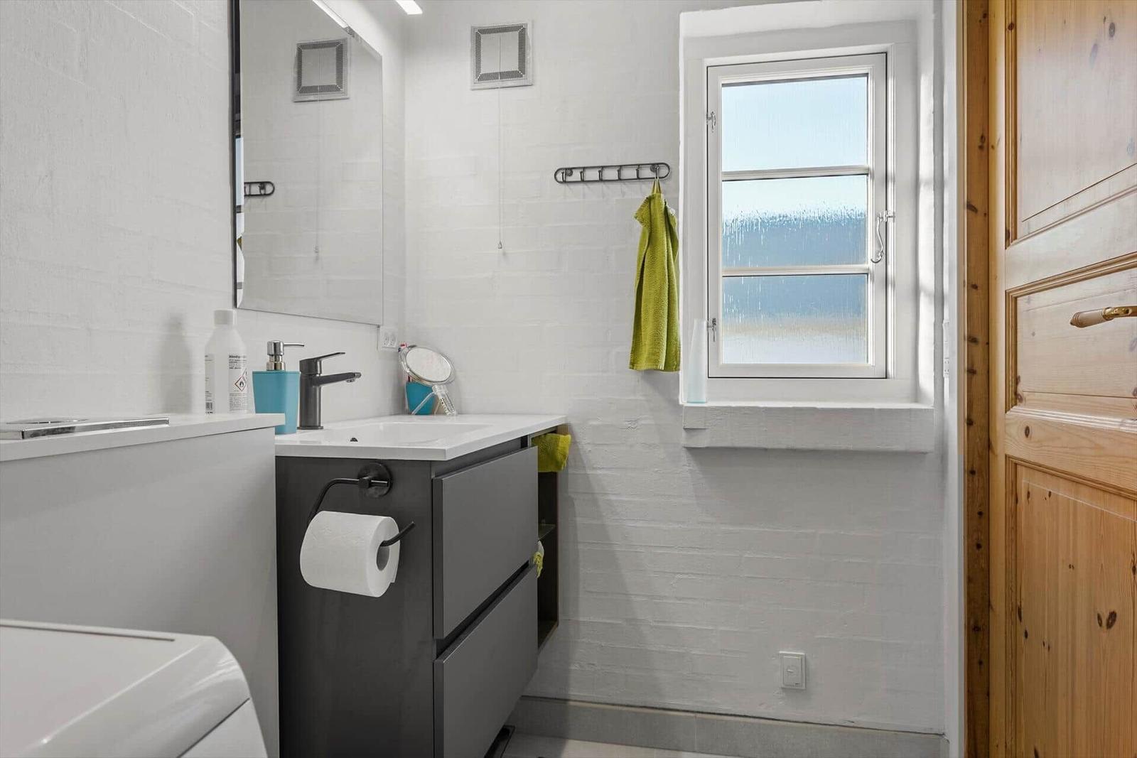 Badezimmer mit Waschbecken, Spiegel und Fenster. Grün Handtuch hängt an Wandhalter.
