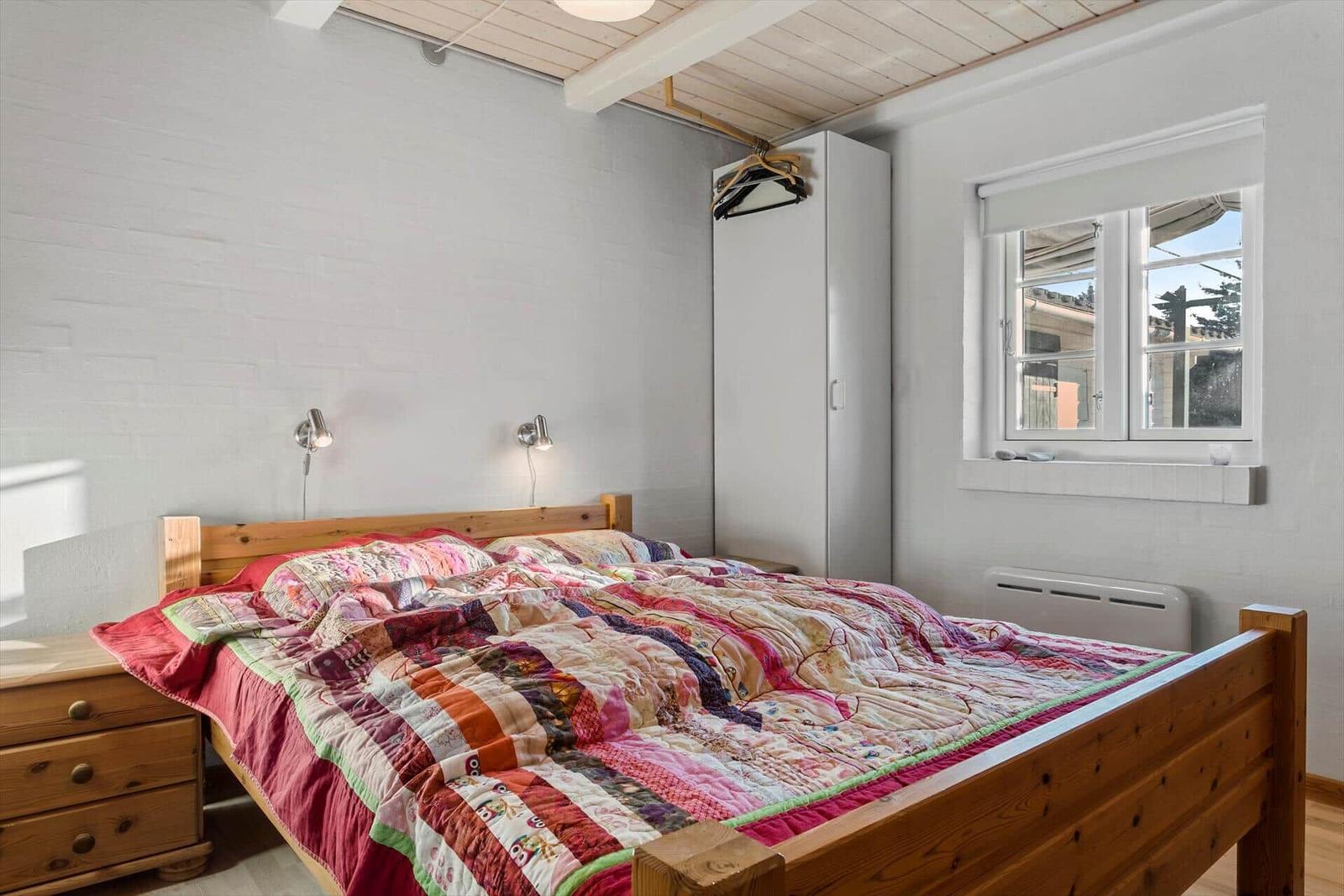 Schlafzimmer mit Holzbett, weißer Wand und Fenster. Kleiderständer und Nachttisch sichtbar.
