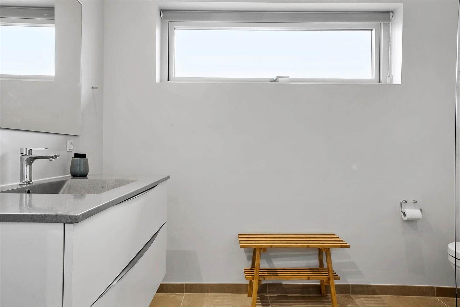 Badezimmer mit Waschbecken, Spiegel, Holzbank und Fenster.