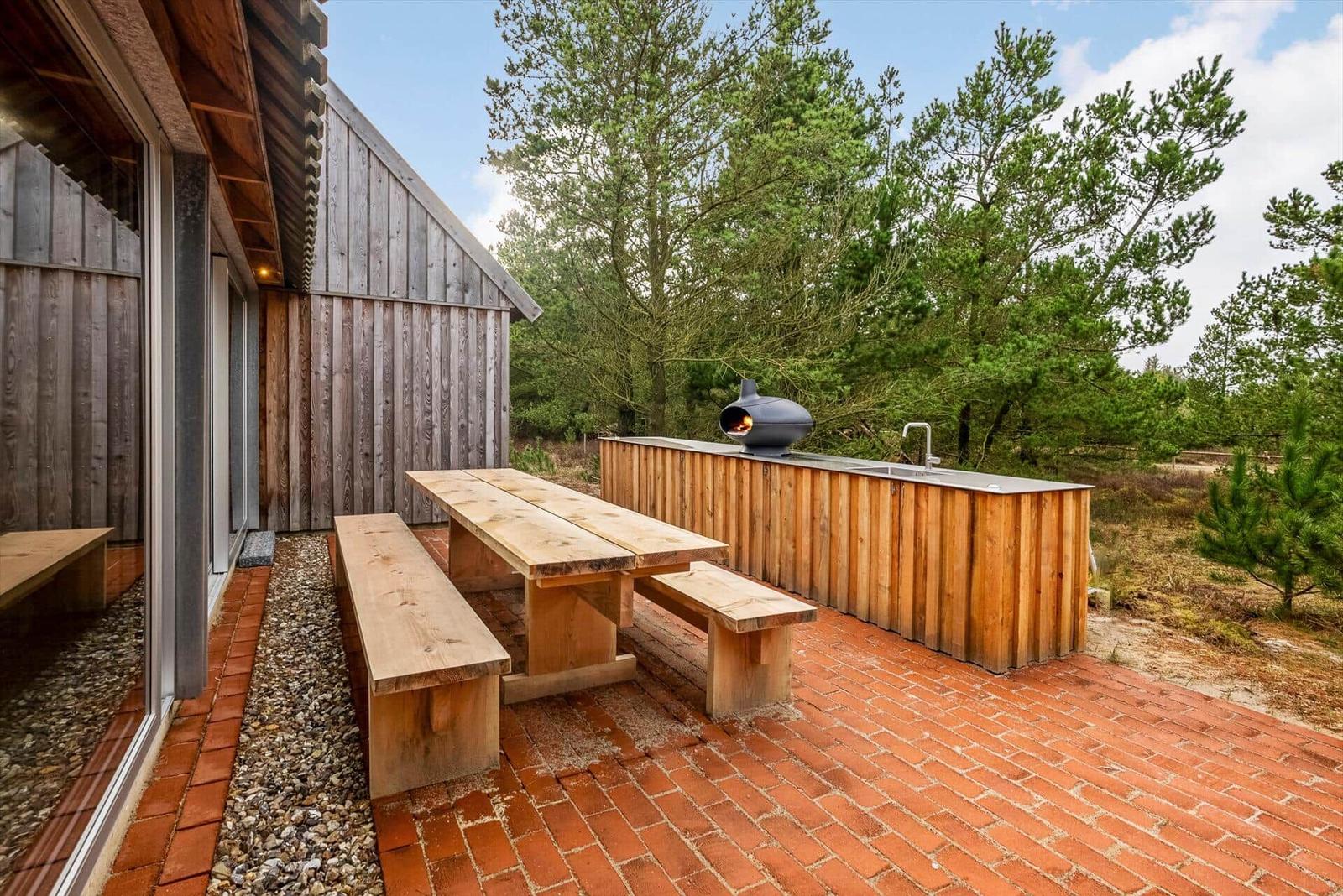 Terrasse mit Holztisch, Banken und Grill. Hintergrund: Bäume und Holzwände.