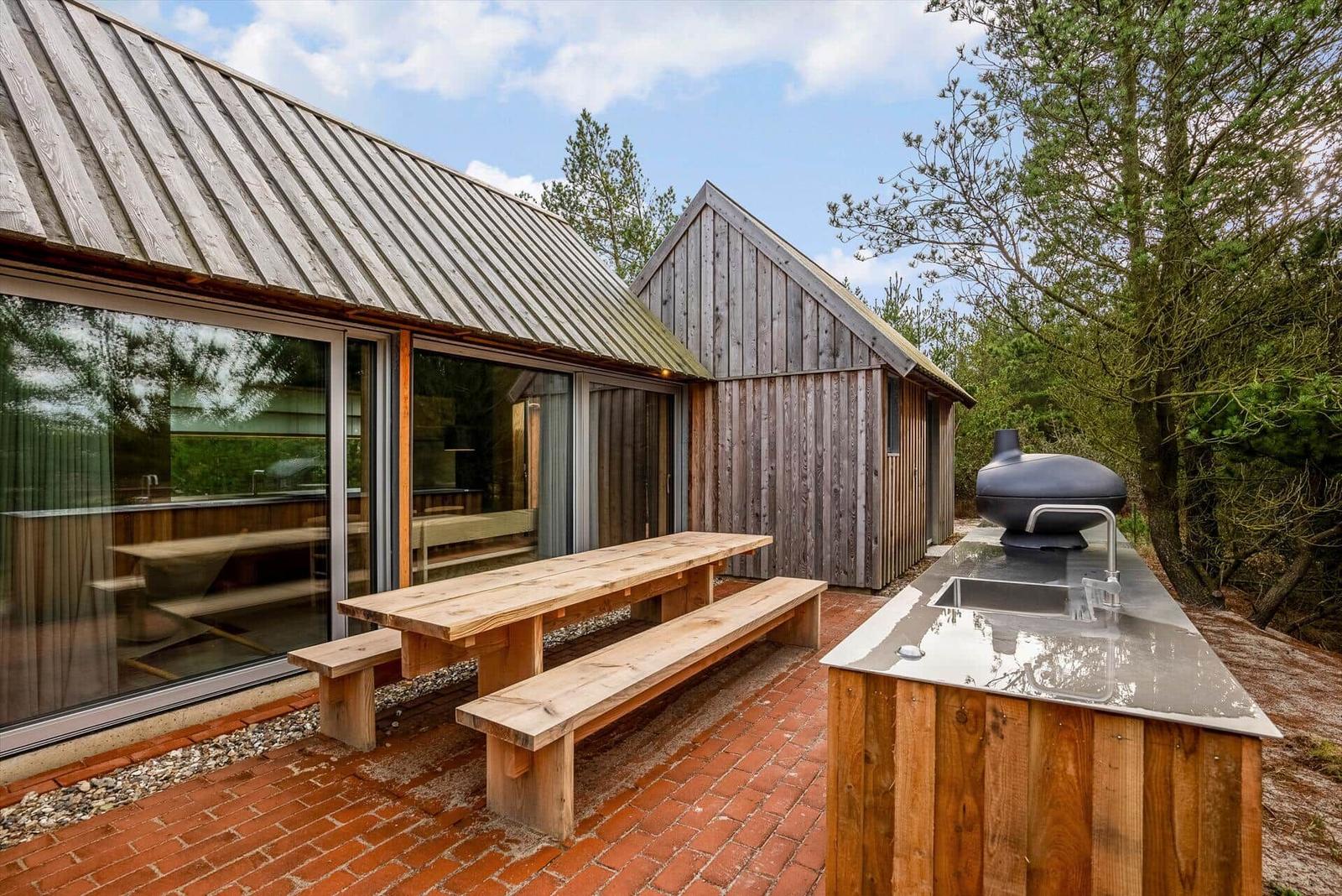 Holzterrasse mit Picknicktisch, Grill und Waschbecken. Glasfront zeigt Inneneinrichtung.