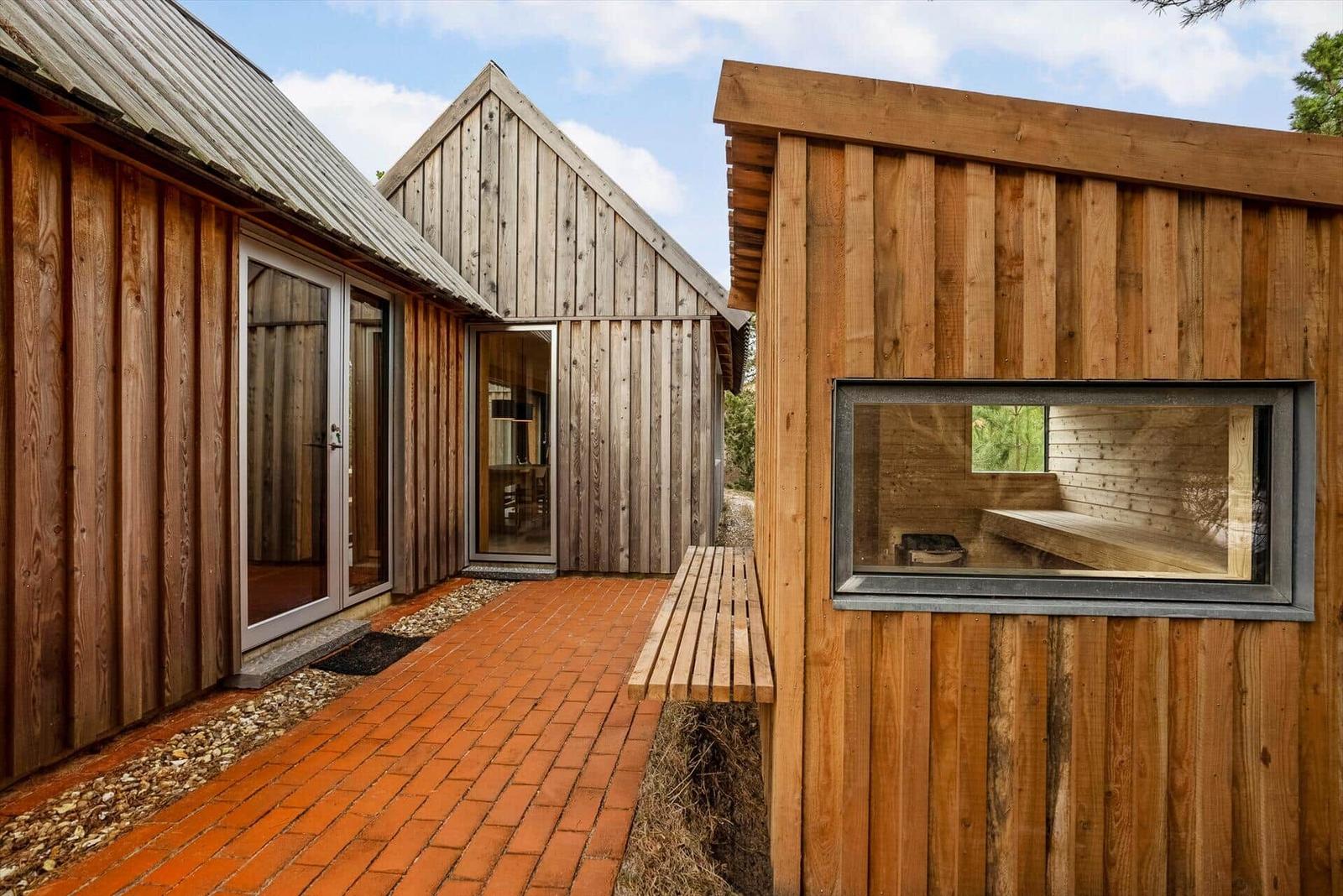 Holzgebäude mit Terrasse und Sauna. Roter Klinkerweg und Fenster zur Sauna.