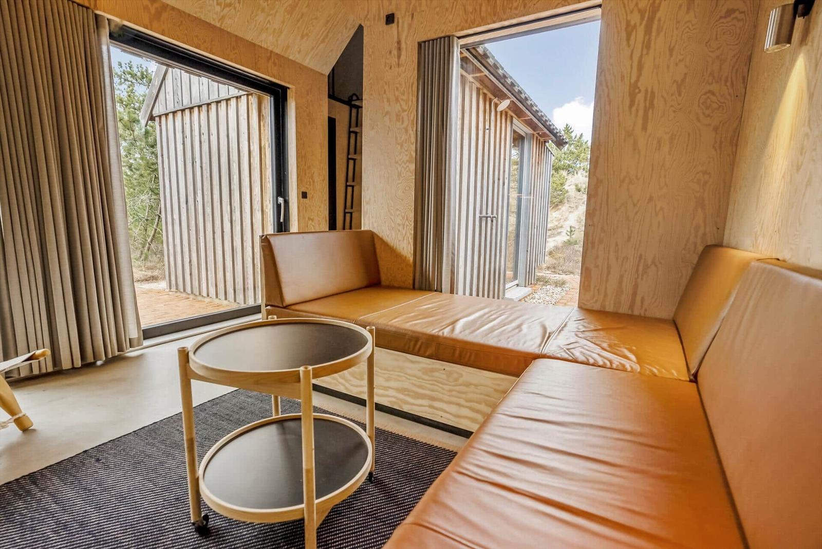 Wohnbereich mit Lederbank, runden Tisch und Blick auf Holzgebäude.