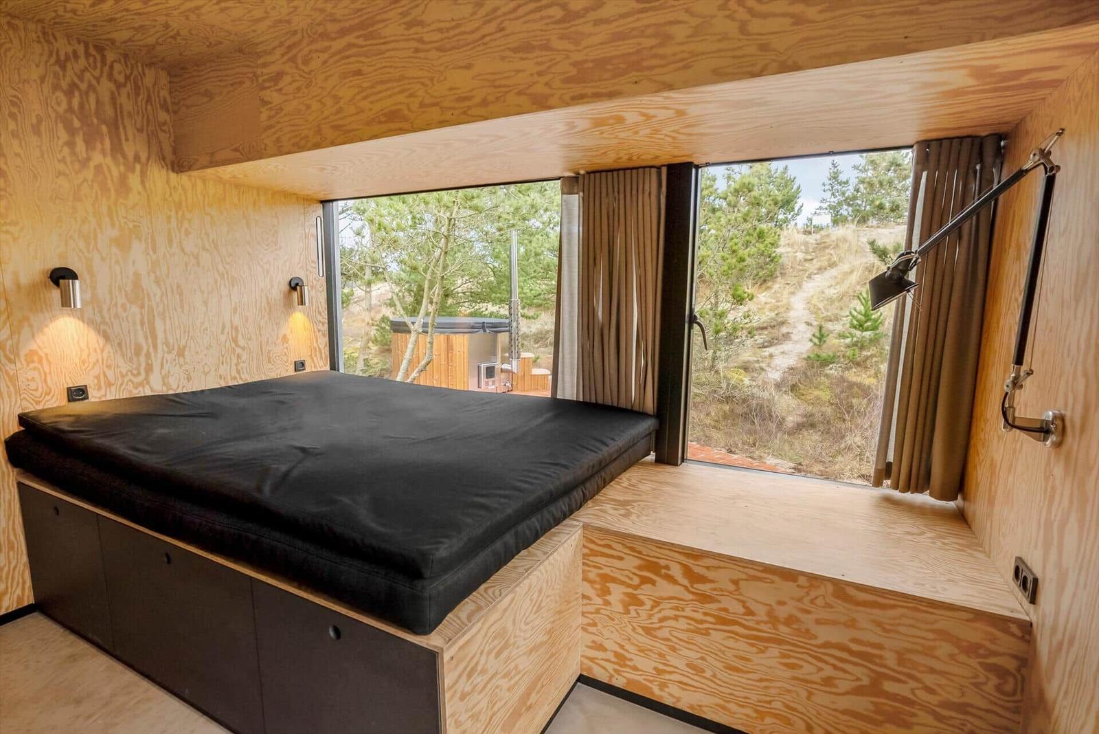Schlafzimmer mit großem Fenster und Holzwandverkleidung. Blick auf Wald und Hügel.