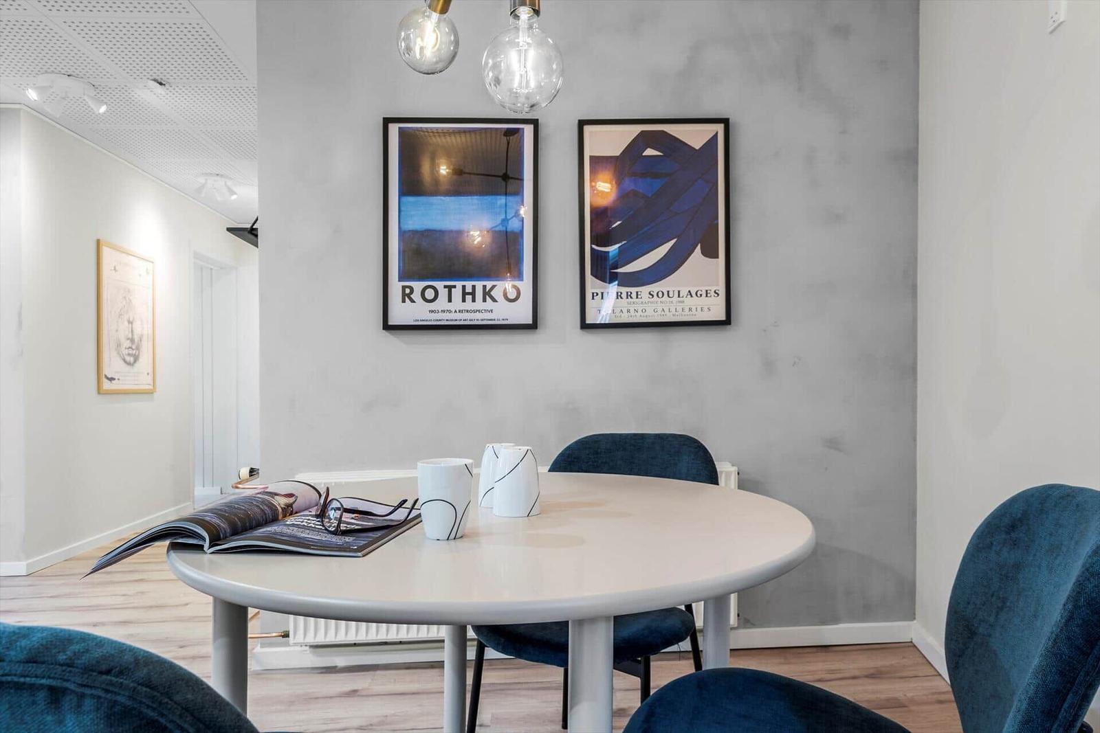 Runde Tisch mit blauen Stühlen, zwei Wandposter und Pendelleuchten.