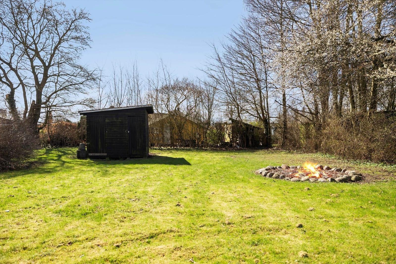 Grüner Garten mit Feuerstelle und kleinem Holzhaus. Bäume umgeben den Außenbereich.