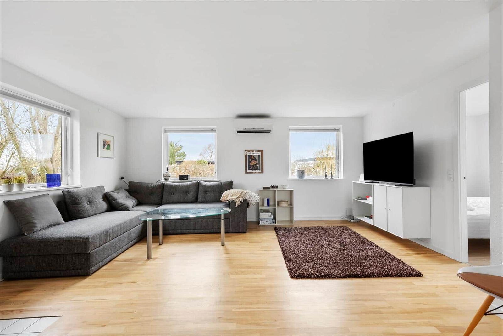Wohnzimmer mit grauem Sofa, Glas-Tisch und braunem Teppich.