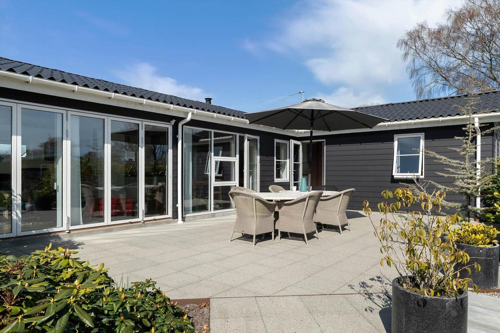 Terrasse mit Tisch, Stühlen und Sonnenschutz vor einem Haus mit Glaswänden.