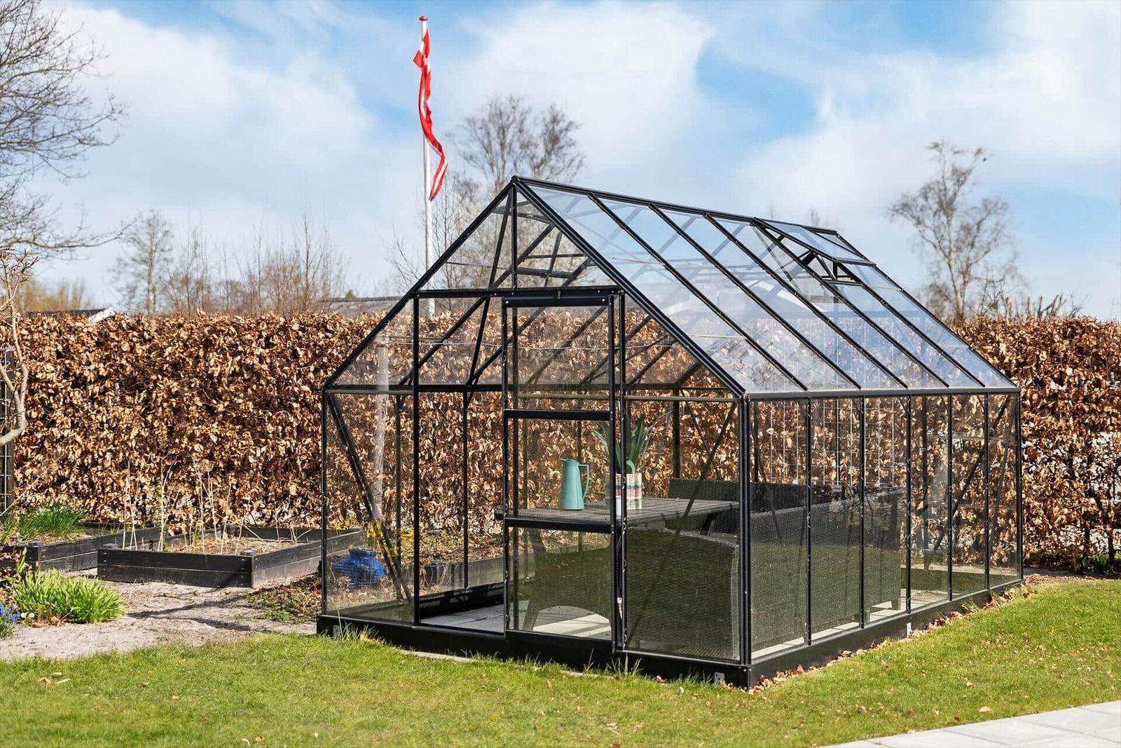 Glasgewächshaus mit schwarzer Metallstruktur im Garten. Hintergrund: Heckenzaun und Flagge.