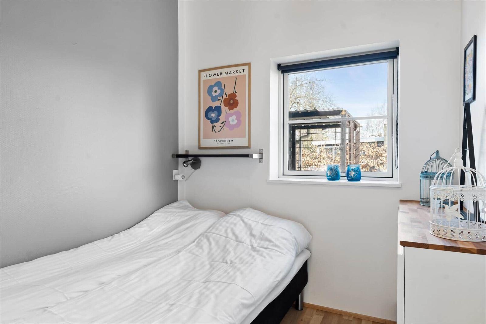 Schlafzimmer mit Bett, Fenster und Wandbild. Auf dem Fensterbrett stehen blaue Vasen.