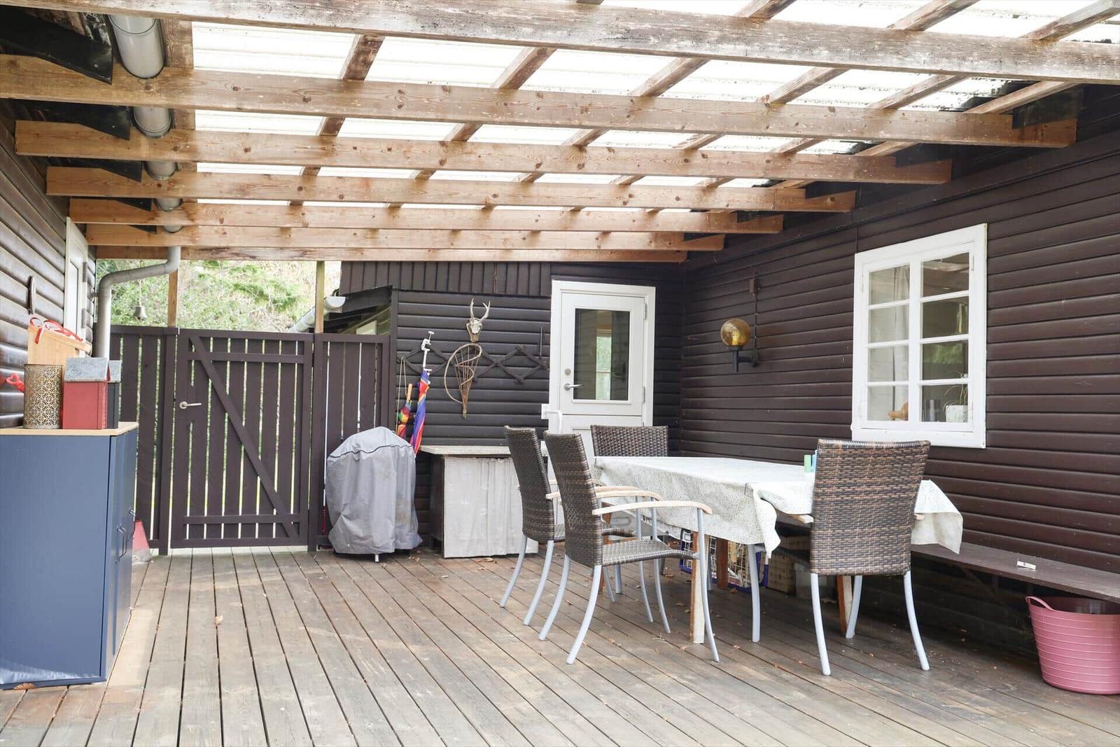 Terrasse mit Holzdecke, Esstisch und Stühlen. Dunkle Holzwand mit Fenster und Tür.
