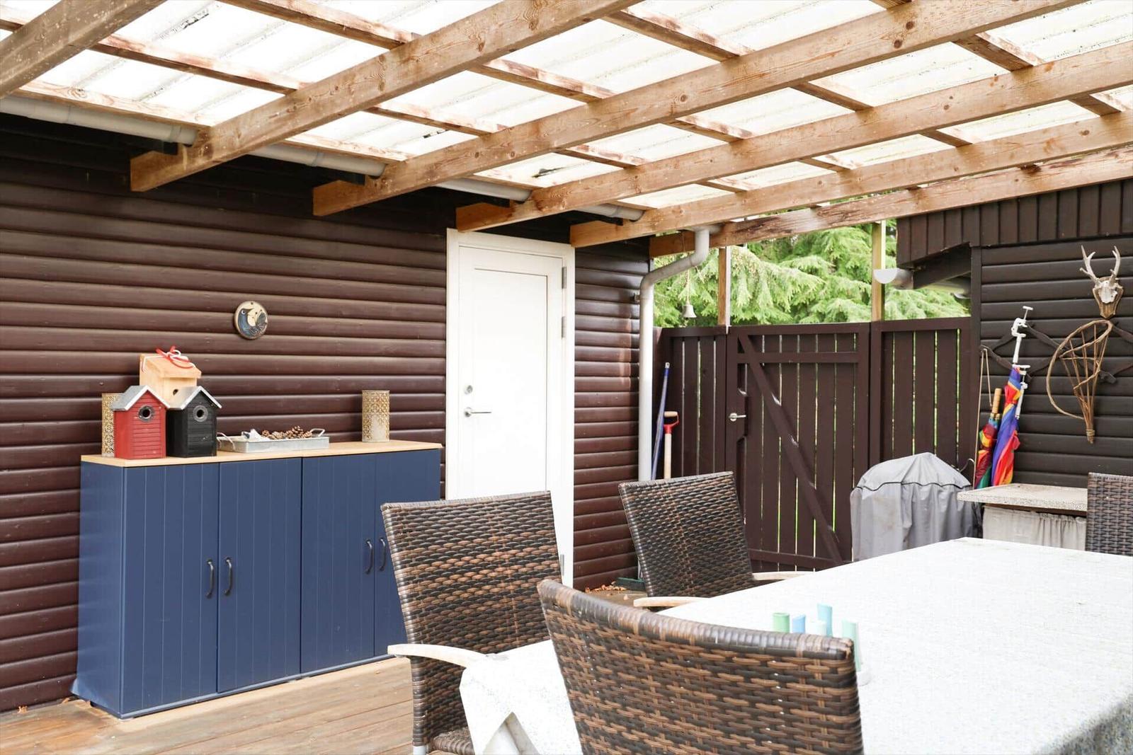 Terrasse mit Tisch, Stühlen und Holzverkleidung. Weiße Tür und Gartenzaun sichtbar.
