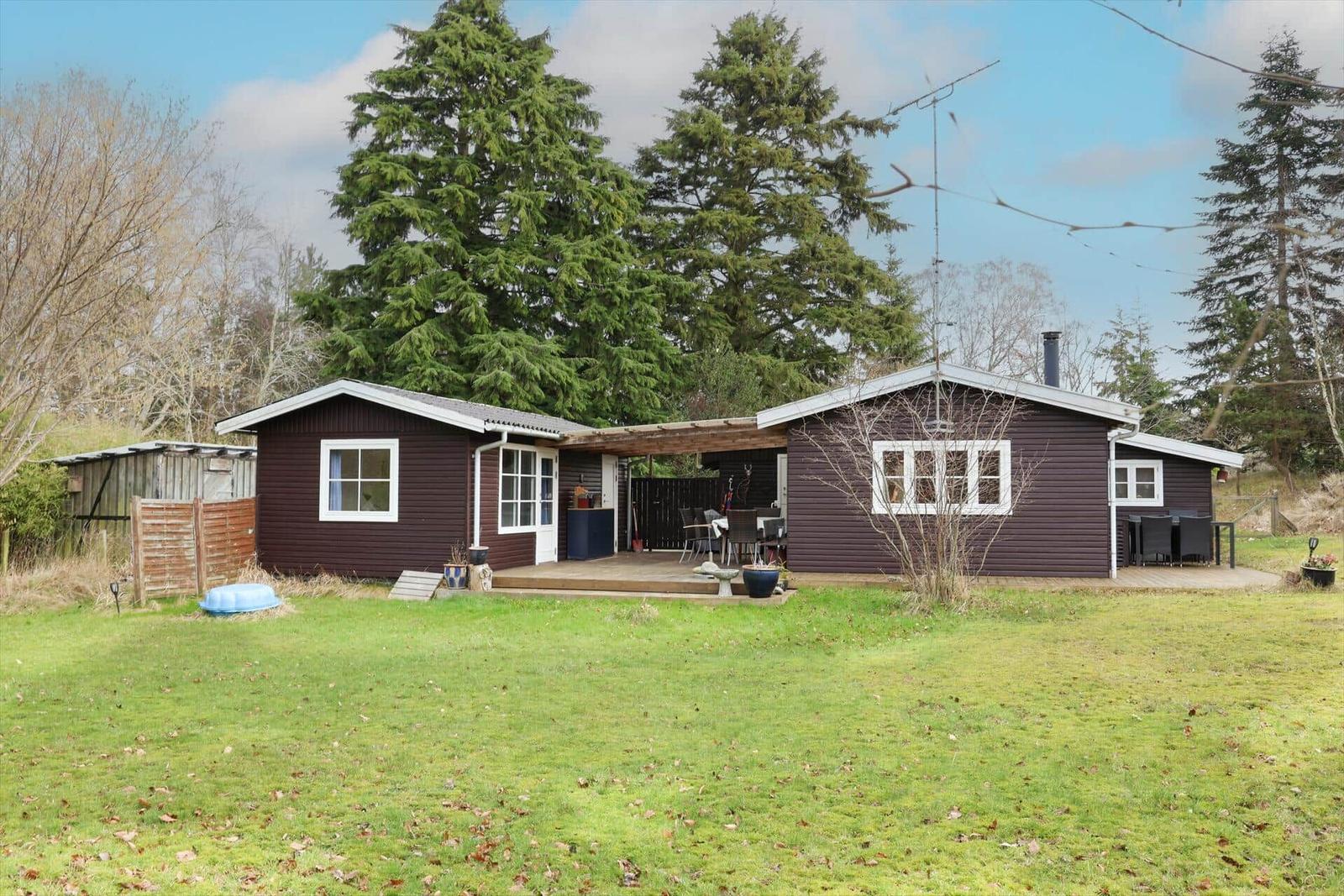 Ein braunes Holzhaus mit Terrasse und Garten. Umgeben von Bäumen und Gras.
