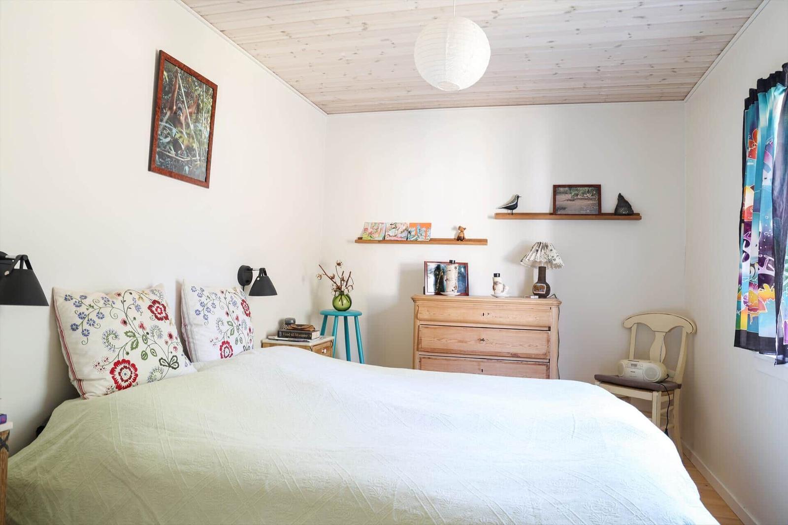 Schlafzimmer mit Bett, Schrank und Wandregal. Decke aus Holz, Fenster mit bunten Vorhängen.
