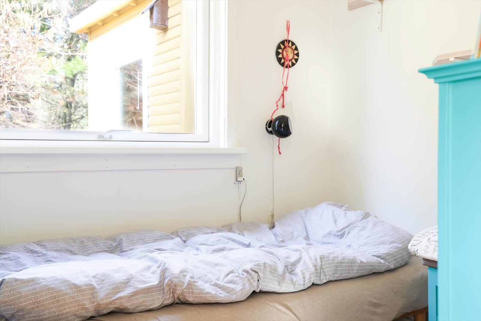 Ein Bett mit gemustertem Bettzeug unter einem Fenster. An der Wand hängt ein Sonnenbild.
