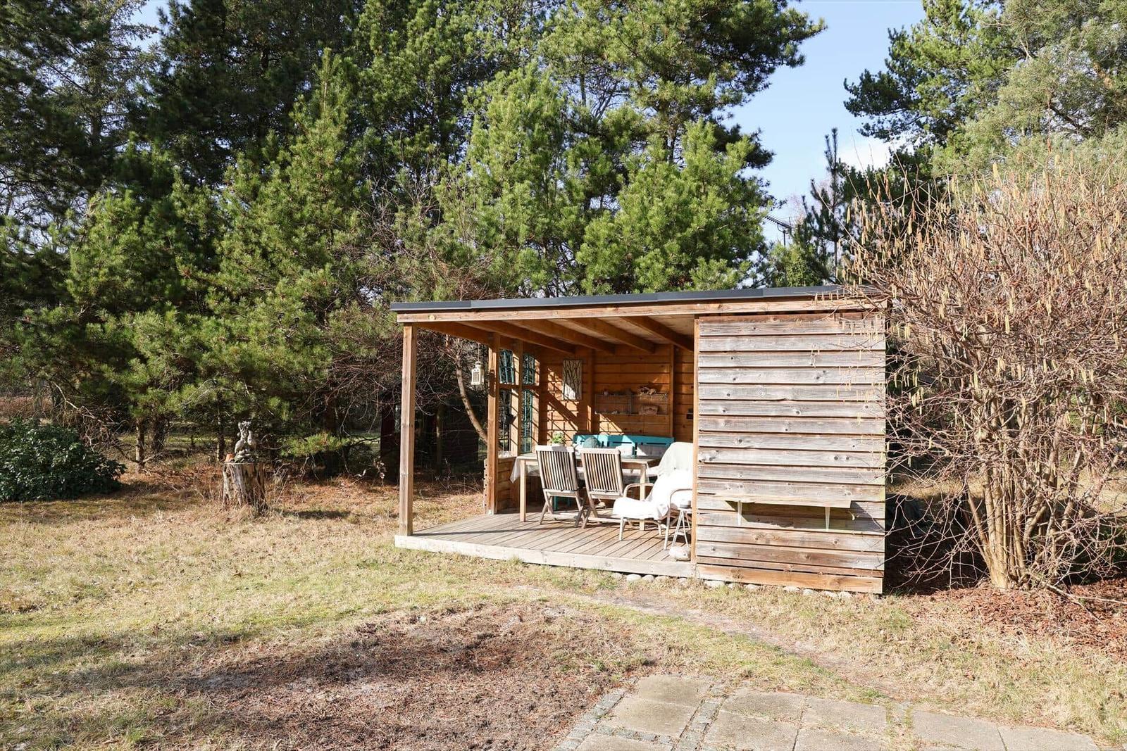 Holzveranda mit Tisch und Stühlen im Freien. Umgeben von Bäumen und Gras.