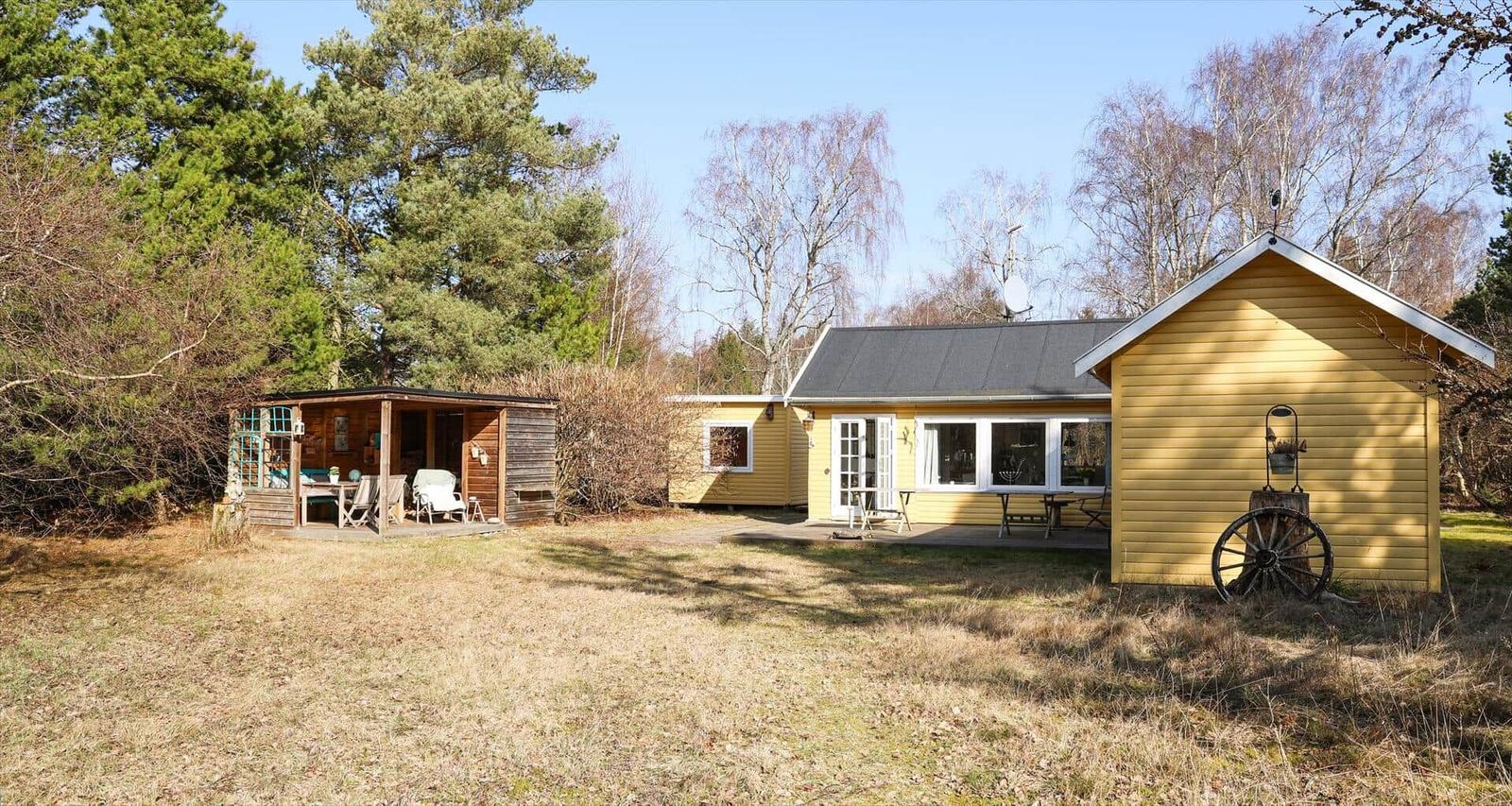 Gelbes Ferienhaus mit Terrasse, Garten und Holzschuppen in Waldnähe.