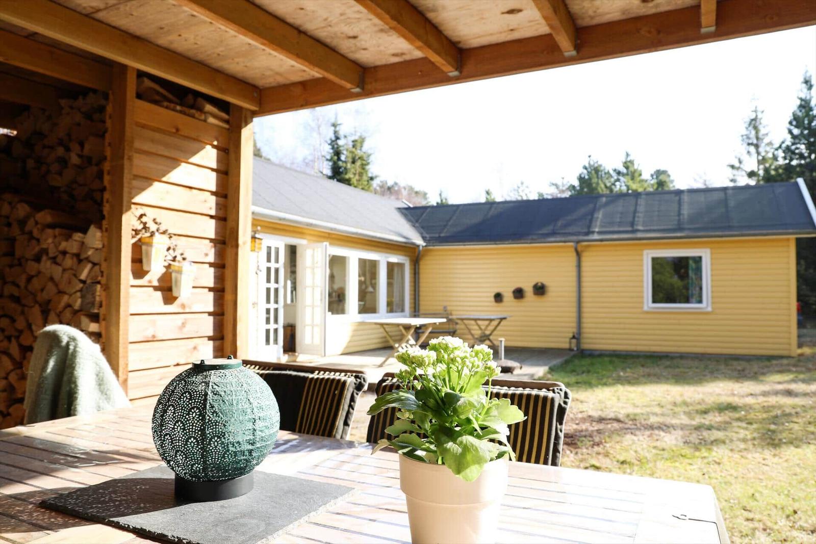 Terrasse mit Tisch, Stühlen und Pflanzen. Hintergrund: gelbes Haus mit Holzveranda und Holzstapel.