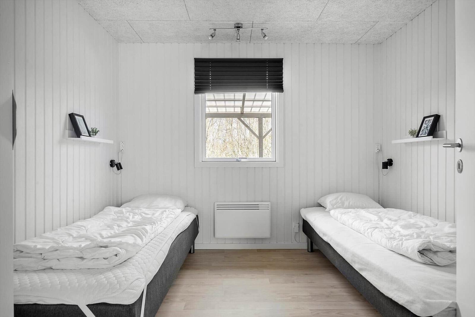 Zwei Einzelbetten, weiß verkleidete Wände, Fenster mit Jalousie, Heizkörper und Wandregale.