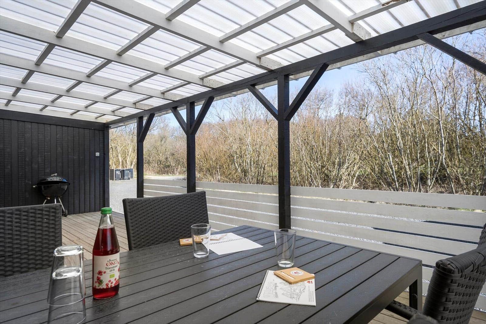 Terrasse mit Tisch, Stühlen und Grill unter Dach. Blick in den Wald.