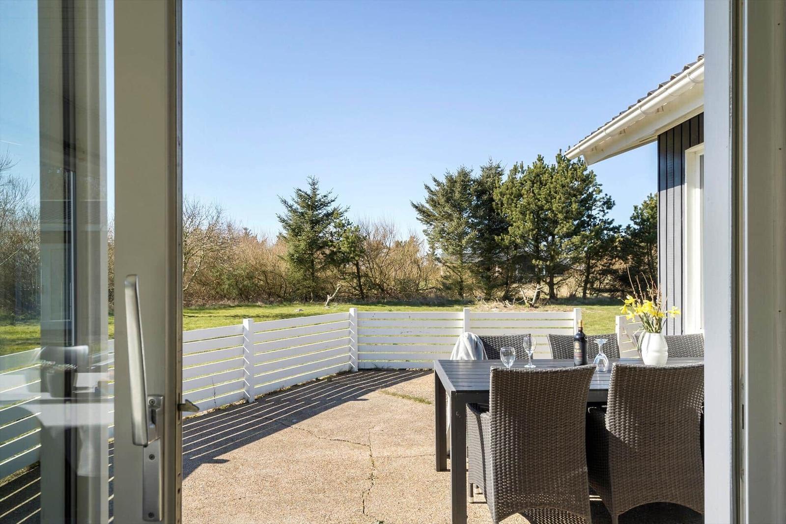Terrasse mit Tisch und Stühlen, weißer Zaun, Grünfläche und Bäume im Hintergrund.