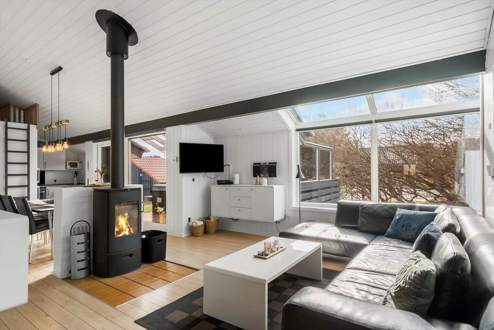 Wohnbereich mit Kamin, Sofa und großen Fenstern. Küche im Hintergrund.
