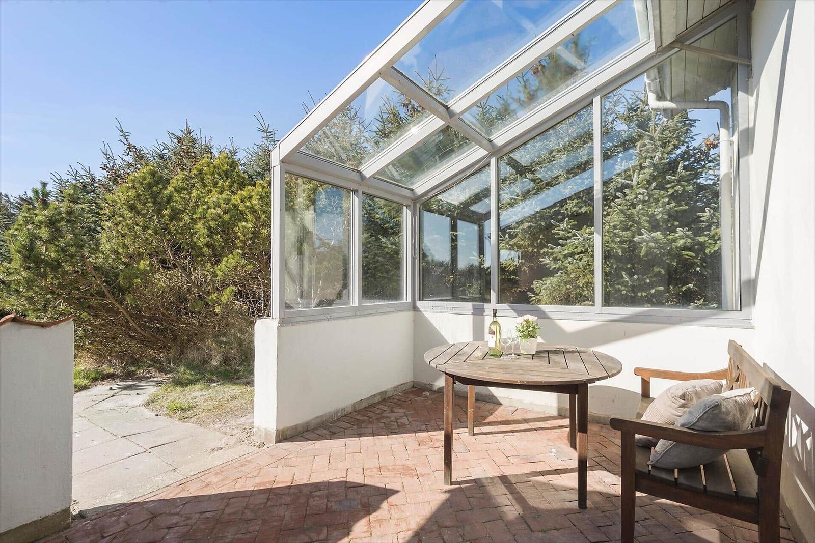 Terrasse mit Glasdach, Holztisch und Bänken. Hintergrund mit Nadelbäumen.