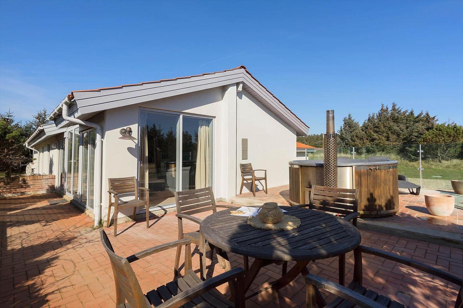 Terrasse mit Holztisch, Stühlen und Whirlpool. Haus mit großem Glasbereich und rotem Ziegelboden.
