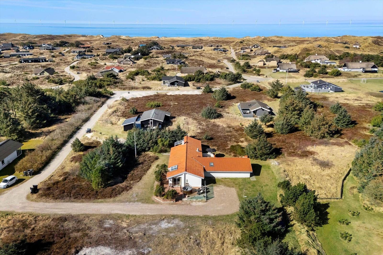 Haus mit orangefarbenem Dach auf einer grünen Wiese, umgeben von Bäumen und Dünen. Blick auf das Meer.