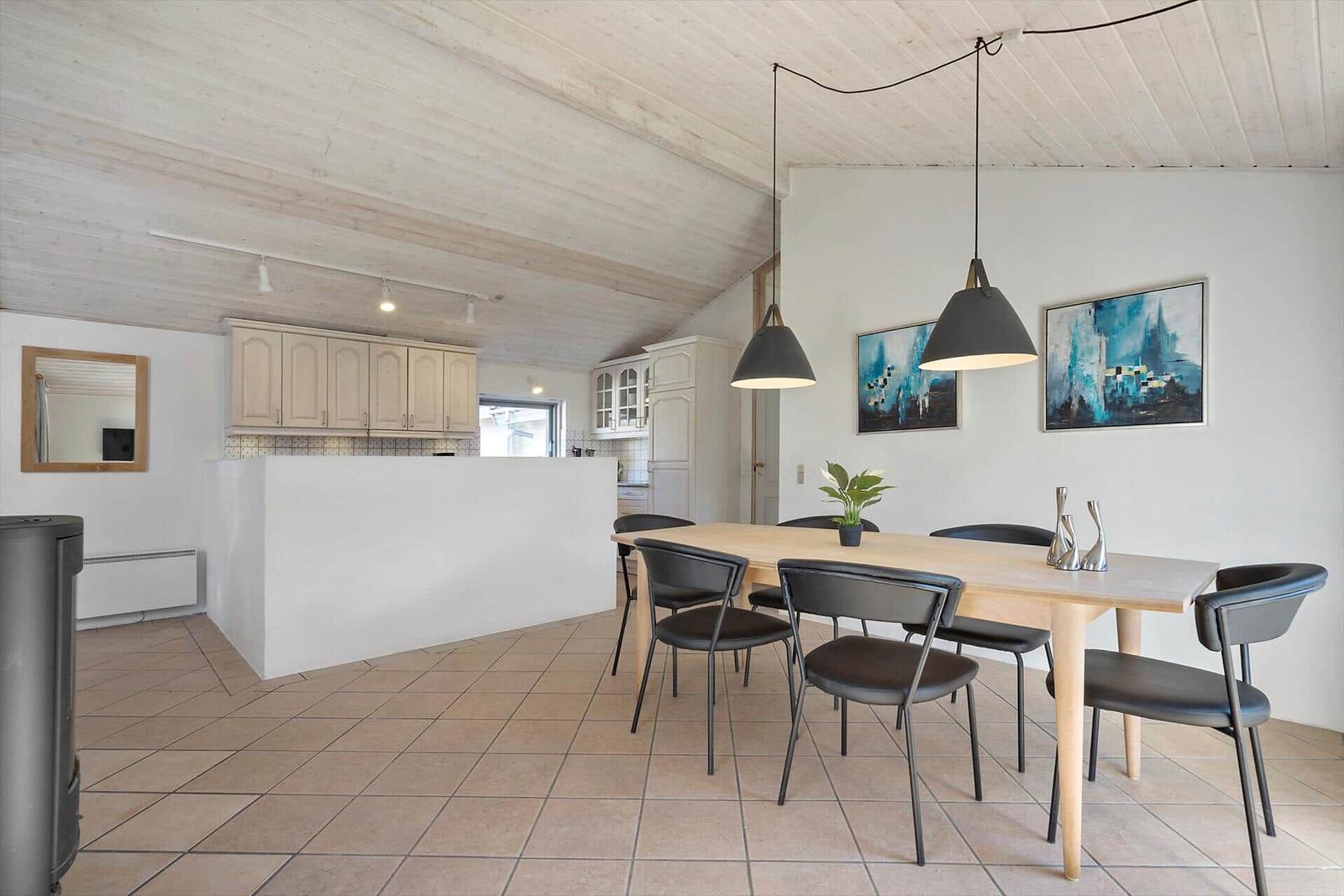 Essbereich mit Tisch und Stühlen unter Hängelampen. Küche mit Schränken und Spiegel. Holzdecke und Fliesenboden.