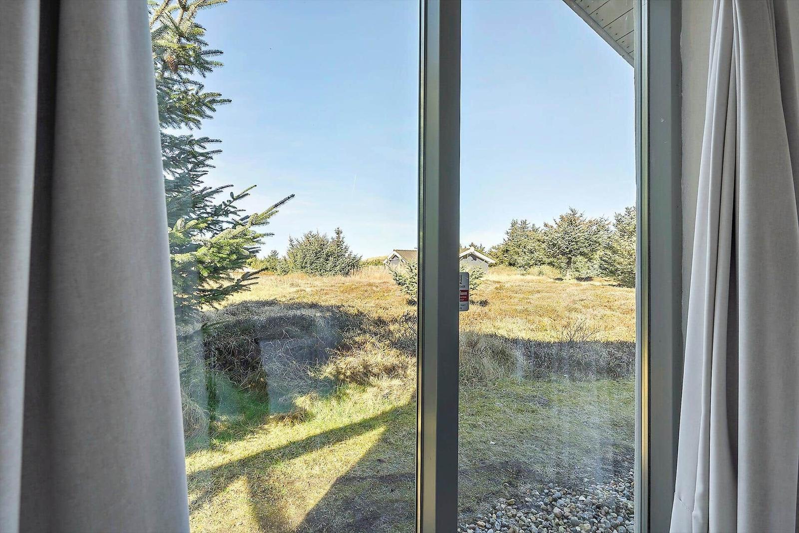 Durch das Fenster blickt man auf einen grasbewachsenen Hang mit Bäumen und einem Haus im Hintergrund.