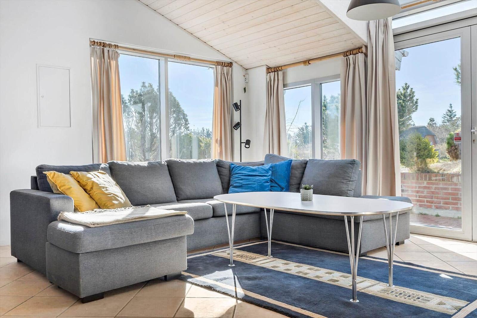 Ein Wohnzimmer mit grauem Sofa, runden Tisch und großen Fenstern.