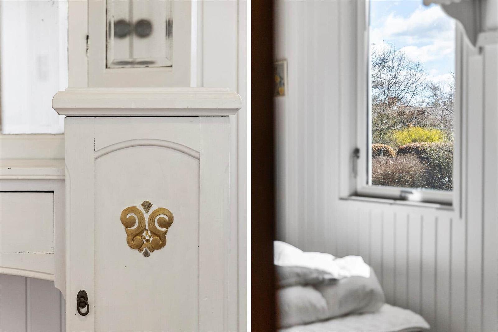 Schlafzimmer mit weißer Holzwand und Fensterblick auf Garten. Antik-Schrank mit goldenem Verzierung.