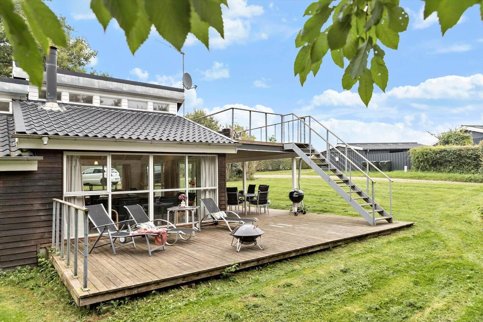 Haus mit Holzterrasse, Gartenmöbeln und Grillplatz unter blauem Himmel.