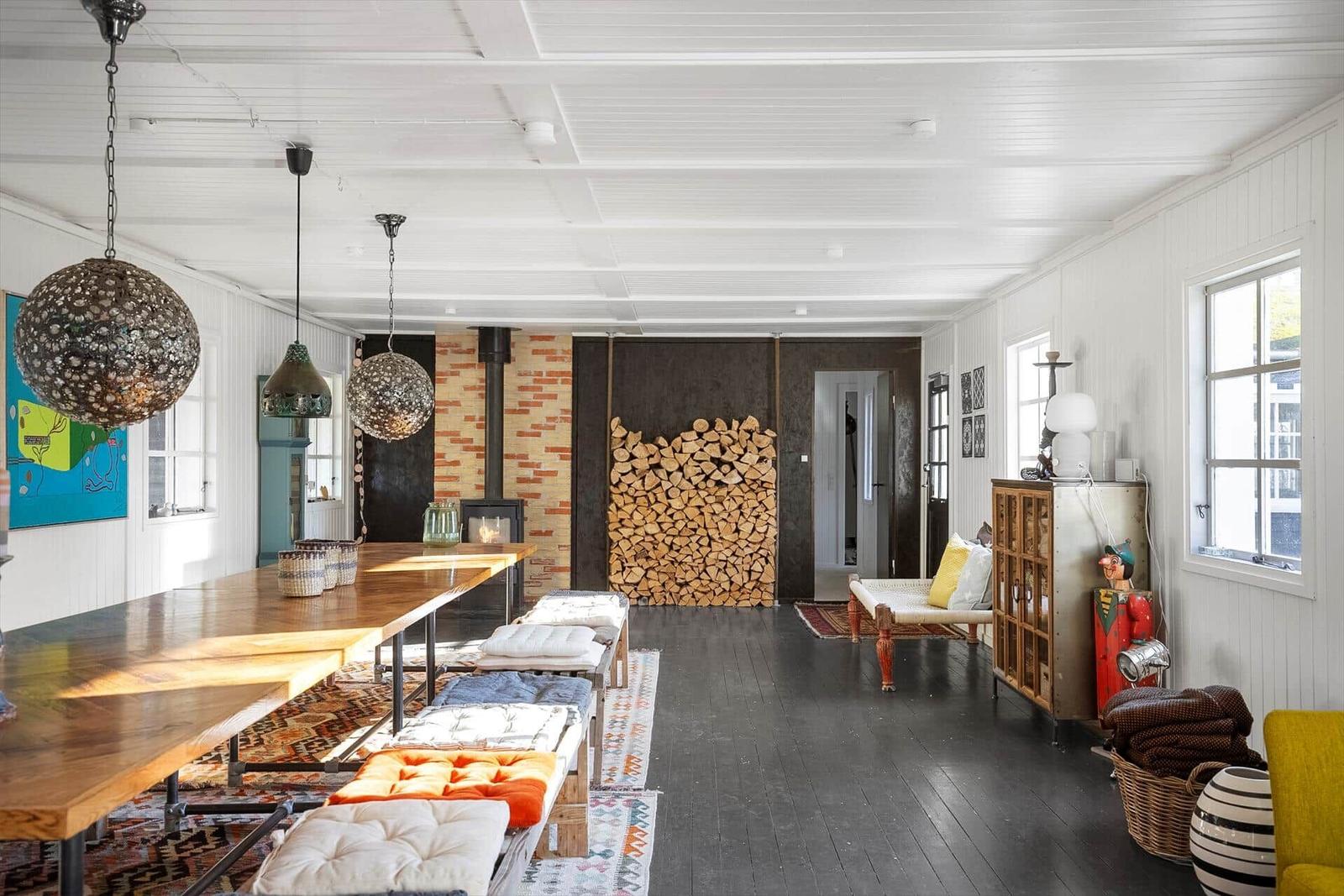 Großer Raum mit Holzfußboden, Tisch, Sitzbänken und Kamin. Holzstapel an Wand. Fenster und Deckenleuchten.