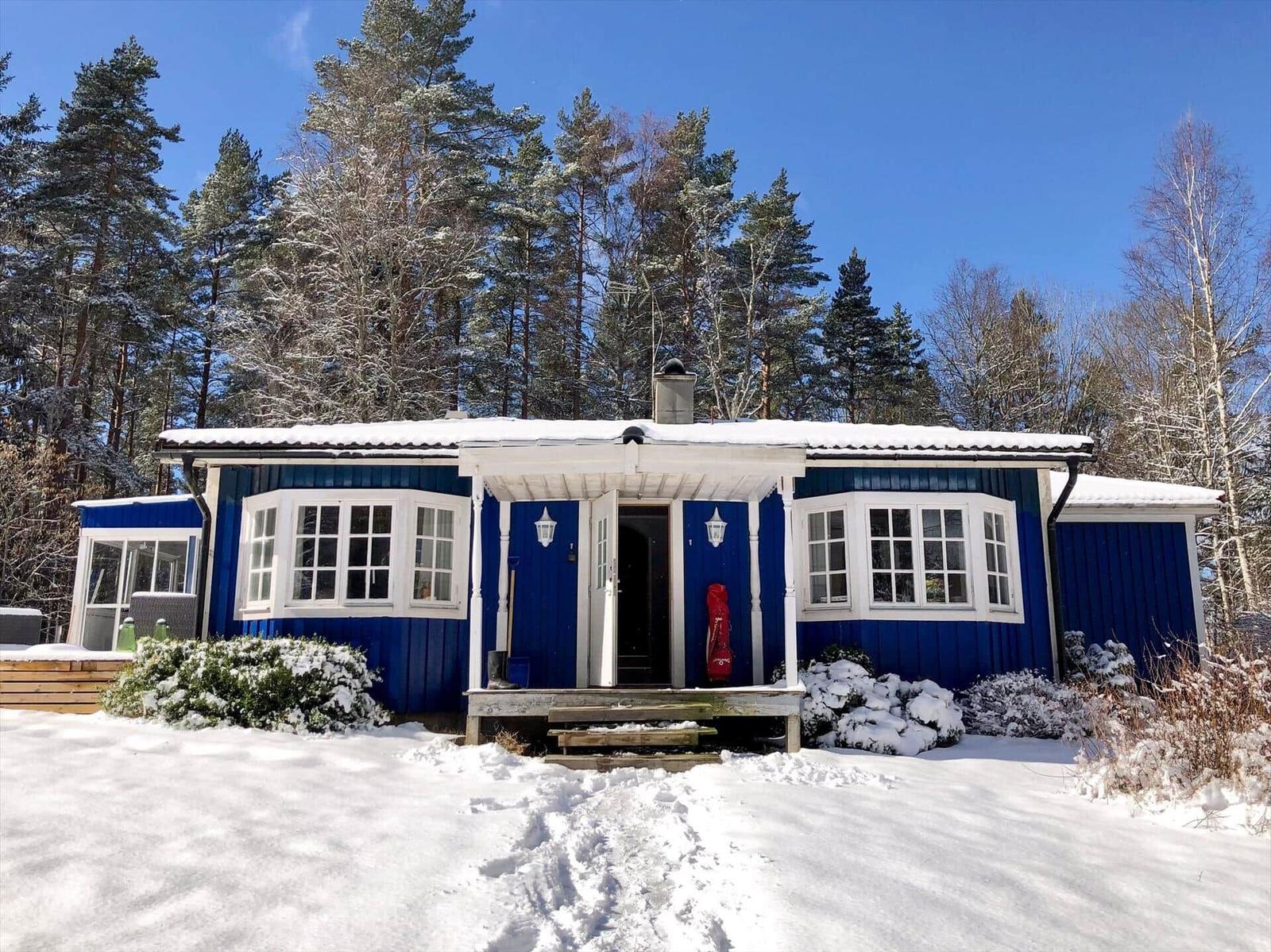 Blauer Holzhaus mit Schnee, weißen Fenstern und Eingangstür im Wald.