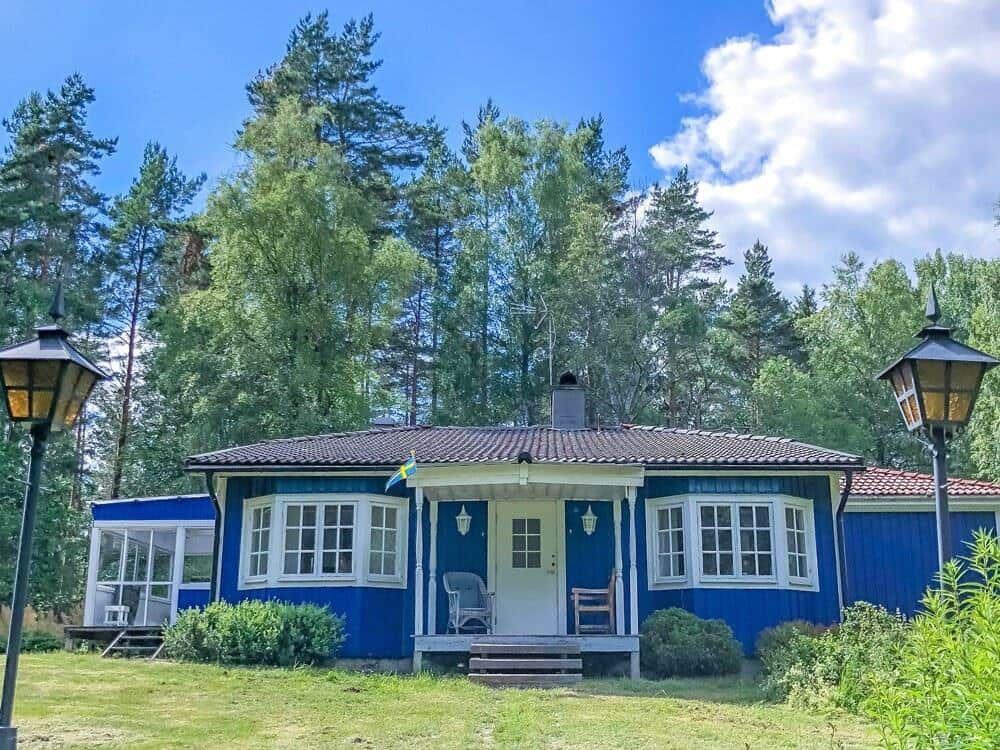 Blauer Holzhaus mit Terrasse und Garten. Umgeben von Bäumen und zwei Laterne.