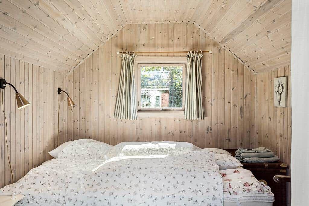 Schlafzimmer mit Holzwand und -decke, Bett mit Blumenbezug und Fenster mit Vorhängen.