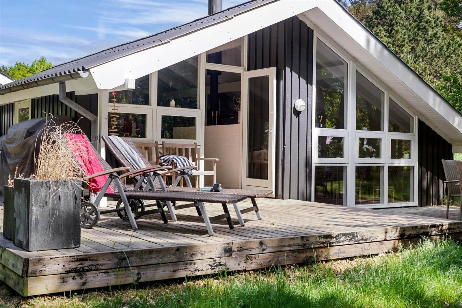 Haus mit Holzterrasse, Liegestühlen und großen Fenstern. Grill und Pflanzen auf der Terrasse.