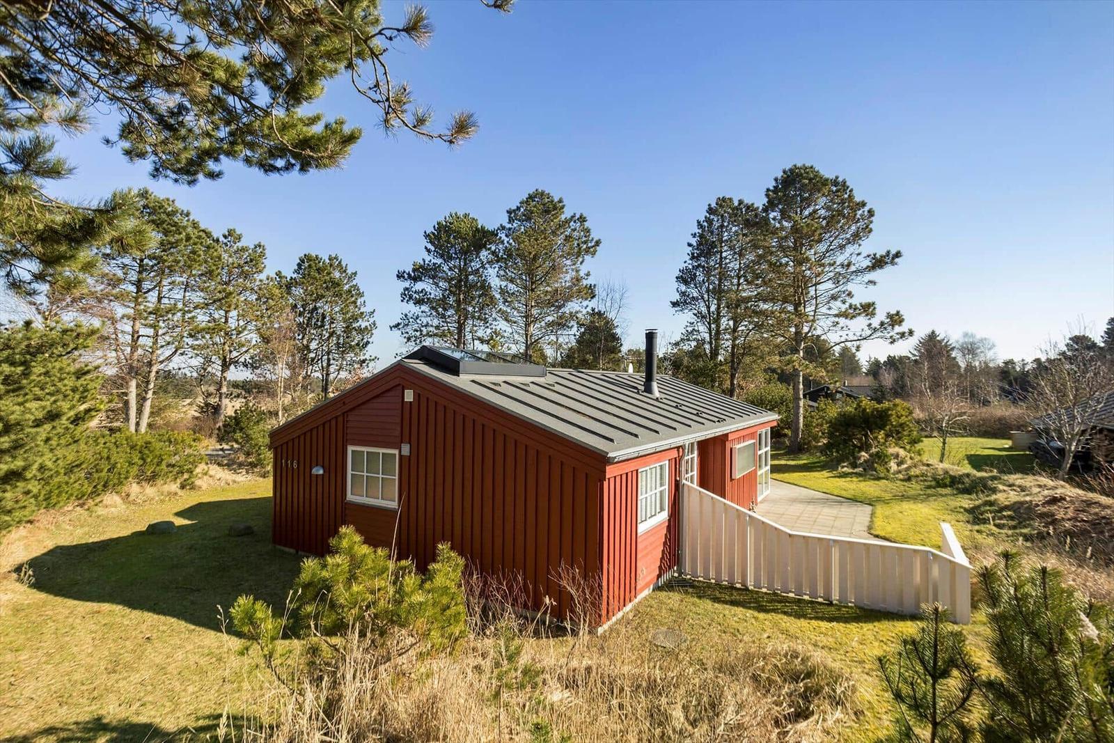 Rotes Holzhaus mit grauem Dach, umgeben von Nadelbäumen und Gras.