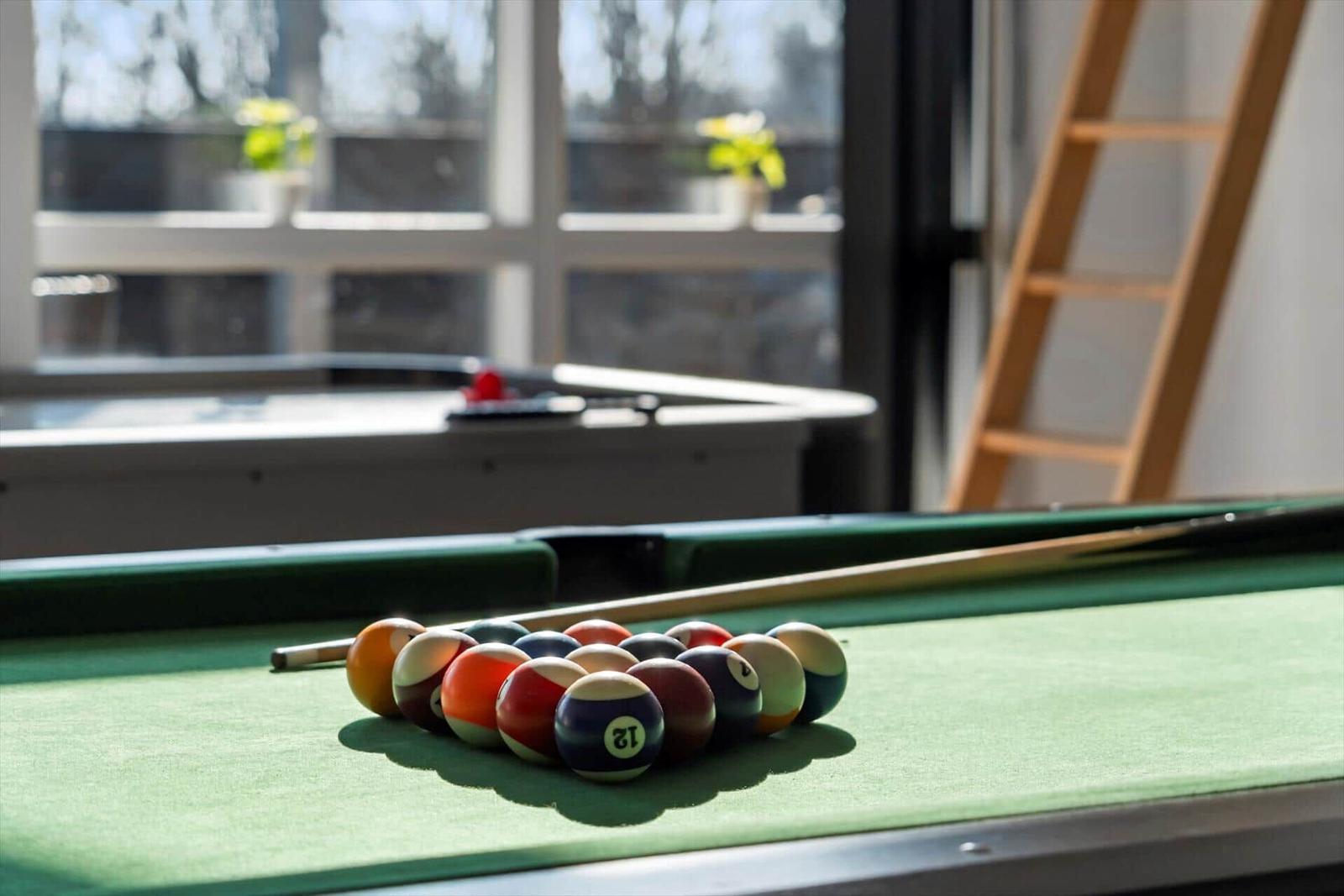 Pool table with balls and cue. In background window and ladder.